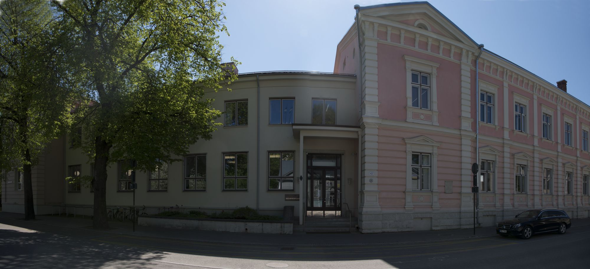 Bänneril on Eesti Kirjandusmuuseumi hoone aadressil Vanemuise 42. Tegemist on panoraamülesvõttega ja maja on näib kaarjana. Paremale poole jääb asutuse vana roosa värviga kaetud majaosa, keskele peauks ja vasakul on valkjas fuajeeakendega majaosa  ja kevadiselt roheliste lehtedega puud.