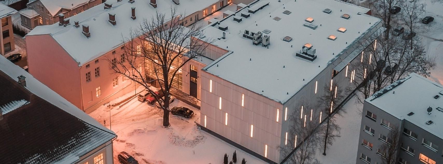 Fotol on droonifoto kirjandusmuuseumi hoone  tagaküljest ülalt. Katused on kaetud õhukese valge lumekihiga. Foto toon on roosakas ja helehall. Akendes tuled põlevad, sest on juba veidi hämar.