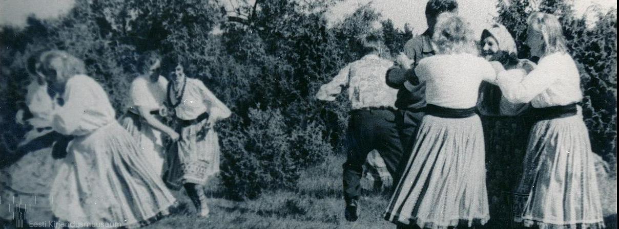 Vanal fotol on näha rahvariietes tantsivad rõõmsad inimesed ja tantsu rõõmsalt pealt vaatavad naised. Taustal on kadakad.